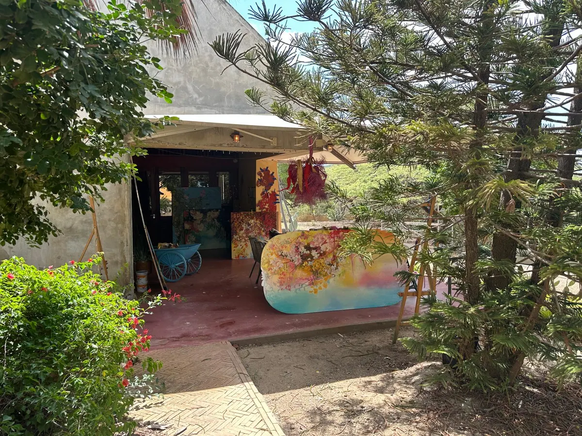 Het atelier van Francis Sling in een tuin op Curaçao: een open deur, een kleurrijk geschilderd doek en bloeiende struiken.